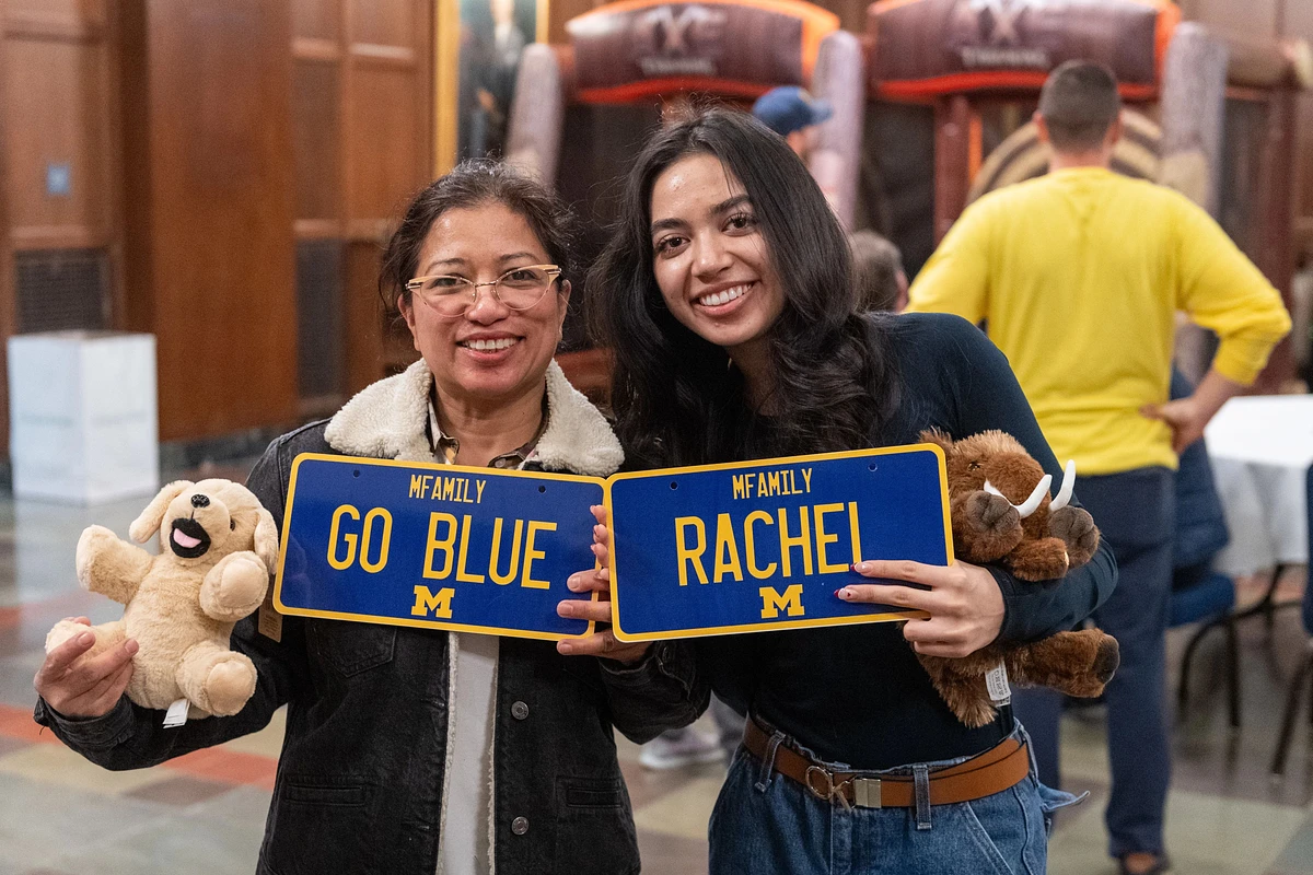 Two women are smiling and holding University of Michigan-themed signs and plush toys at an indoor event.