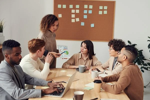 A group of six people are having a meeting at a table, with colorful sticky notes on a board in the background.