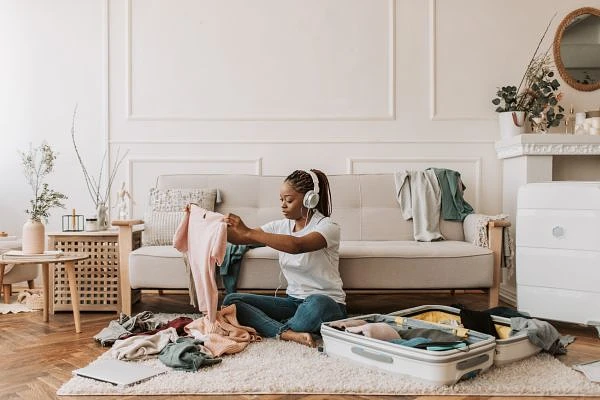 A person sitting on the floor is packing clothes into a suitcase, wearing headphones in a cozy living room.